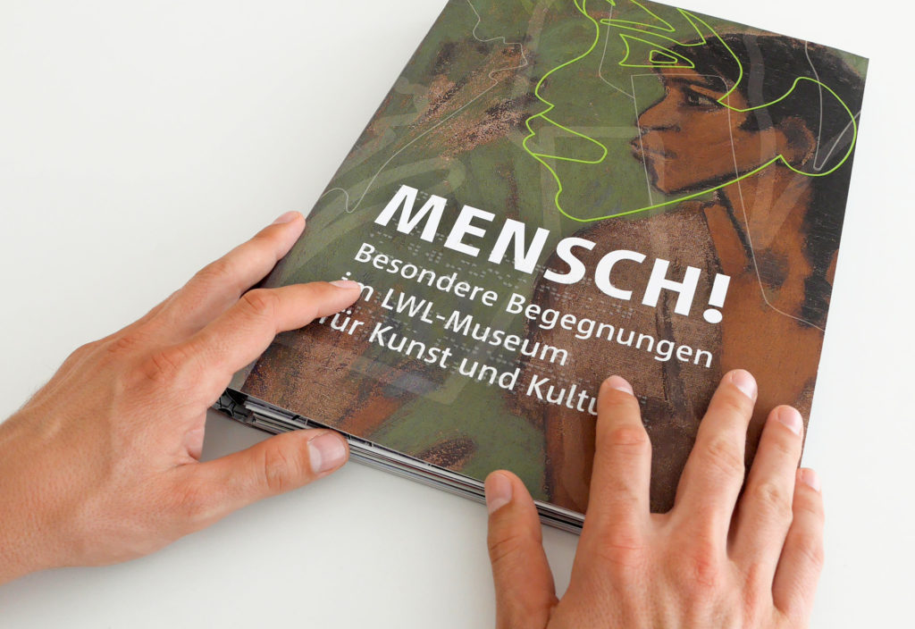 Auf dem Projektbild ist das tastbare Cover des Museumsführers mit der Aufschrift: „Mensch! Besondere Begegnungen im LWL-Museum für Kunst und Kultur“ und Braille-Schrift.