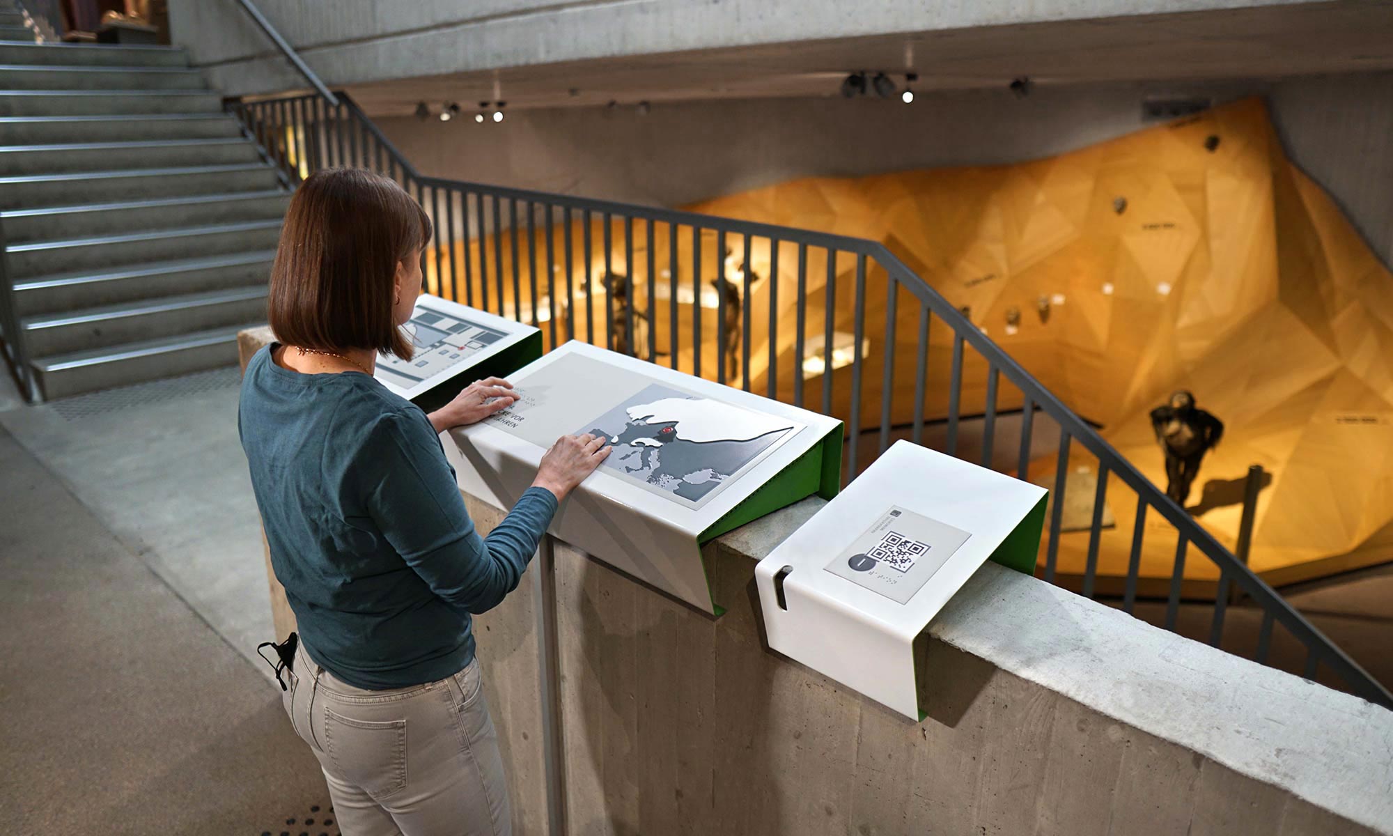 Tamara Ströter stands at a tactile model on which a map of Europe is depicted. This tactile model is a curved shape made of sheet steel, which is mounted on a wall offset at a slight angle. Behind the wall is a staircase that leads to other floors of the museum. Looking out over the banister, one has a view of parts of the exhibition on the lower floor.