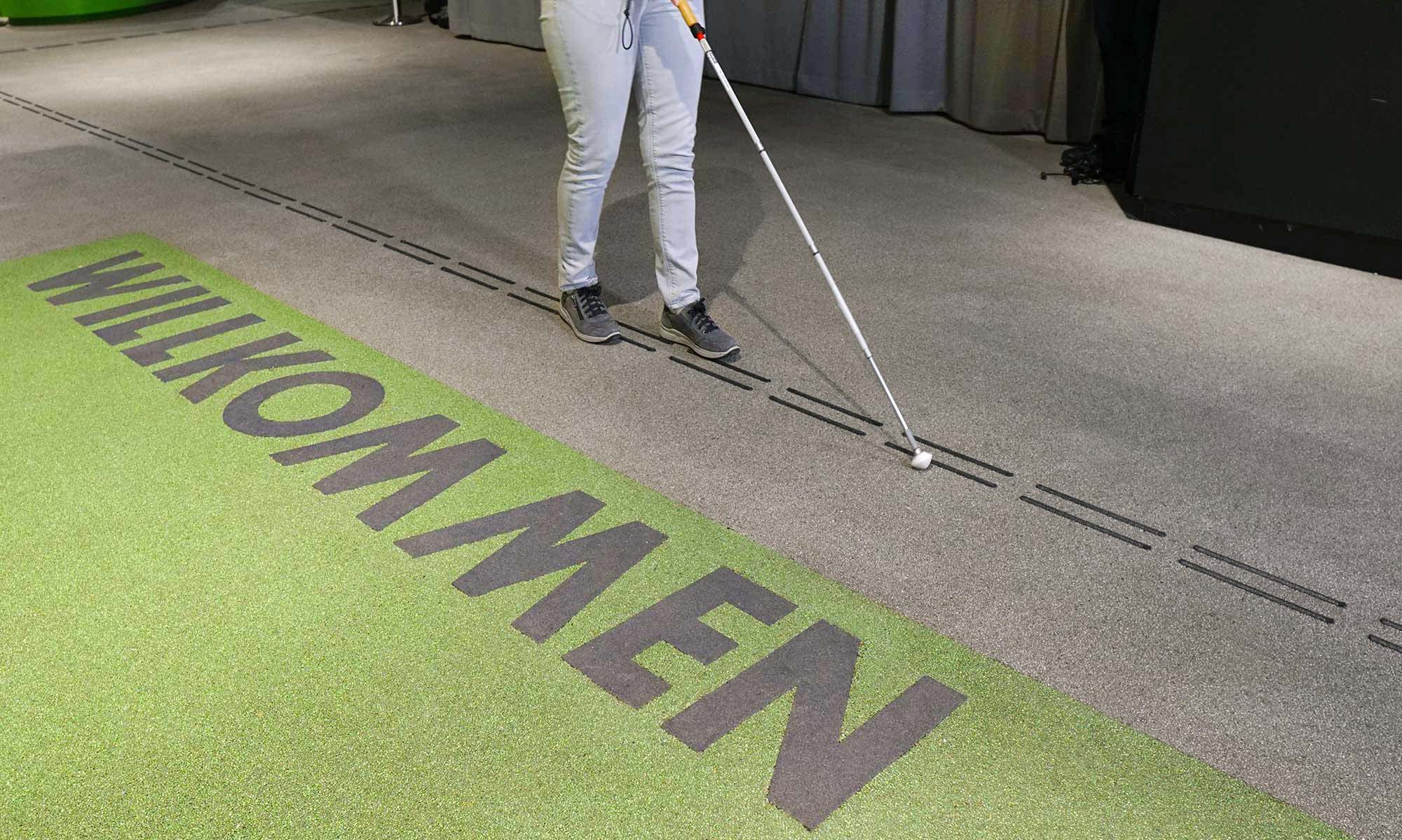 On the picture you can see floor indicators in the entrance area of the museum. A straight path is shown with two adjacent dashed black lines. These go right across the picture. There is a visitor walking along the markings with her cane. In the foreground, there is a large green area on the floor that reads: Welcome.