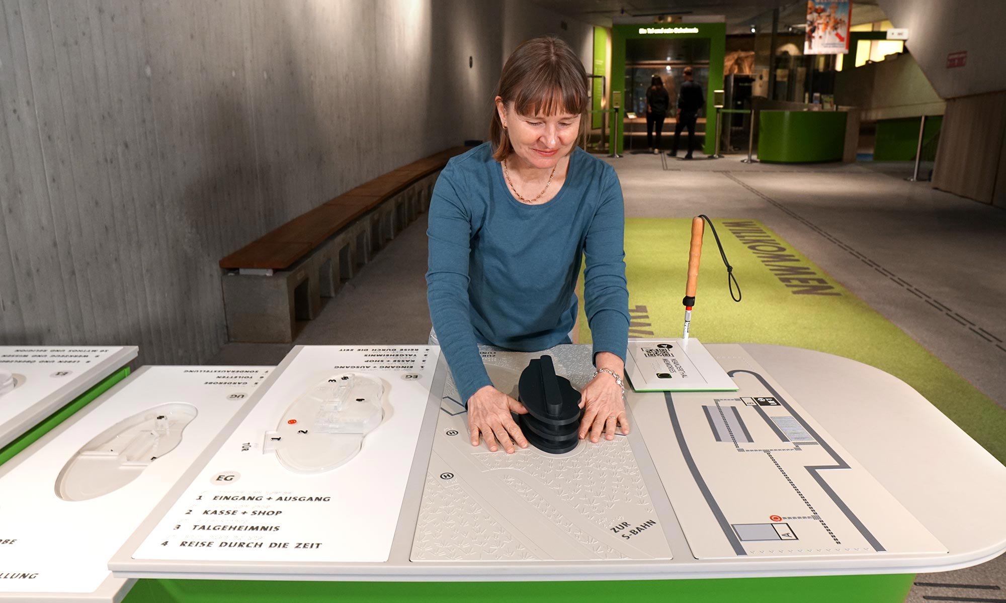 Tamara is standing at a large table designed by inkl.Design in the entrance area of the Neanderthal Museum. On the table are several tactile map of the individual floors of the museum. She feels the dimensions of the building on the basis of a 3D model, which is also attached to the table. In the background there is a view of visitors leaving the building...