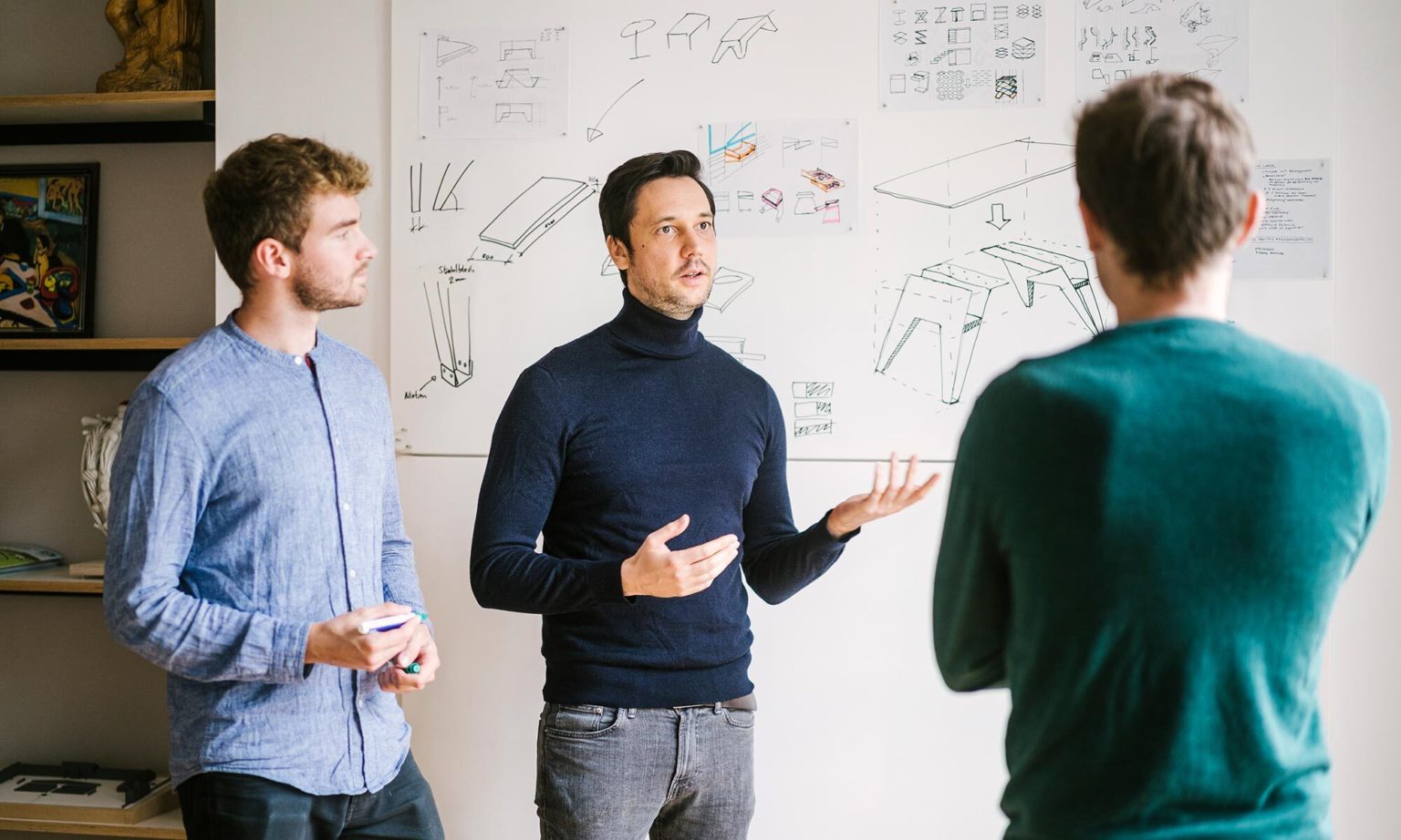 Several product designers from inkl.Design stand in front of a whiteboard showing sketches of a tactile table and exchange ideas.