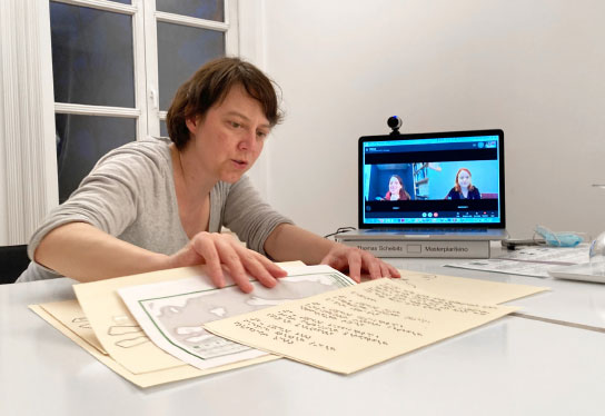 The picture shows a focus group meeting where some members are connected via video conference. The incl.design employee Franziska Müller sits at a table and picks out something in her documents during the conversation.