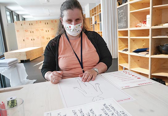 A blind woman from the focus group is sitting at a table. In front of her is a poster from the Body Cartographer. She feels the tactile outline of the person on the poster. Despite her mask, you can see that she is smiling.