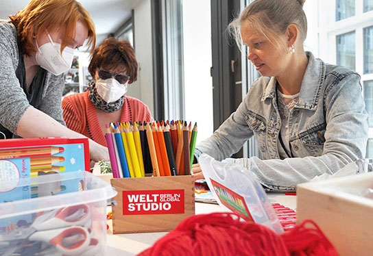 The photo shows three women from the focus group sitting at a table. In the foreground are piles of colourful pens, boxes of craft materials and coloured yarn. The pencil box is labelled with a sticker from the Weltstudio, white writing on a red background.