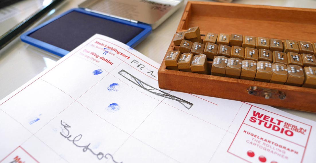 The photo shows a close-up of small stamps marked with tactile braille letters, next to an ink pad. In the foreground is a piece of paper with the logo of the Weltstudio, which belongs to the Rolling Cartographer. On it, visitors can describe their favourite place.