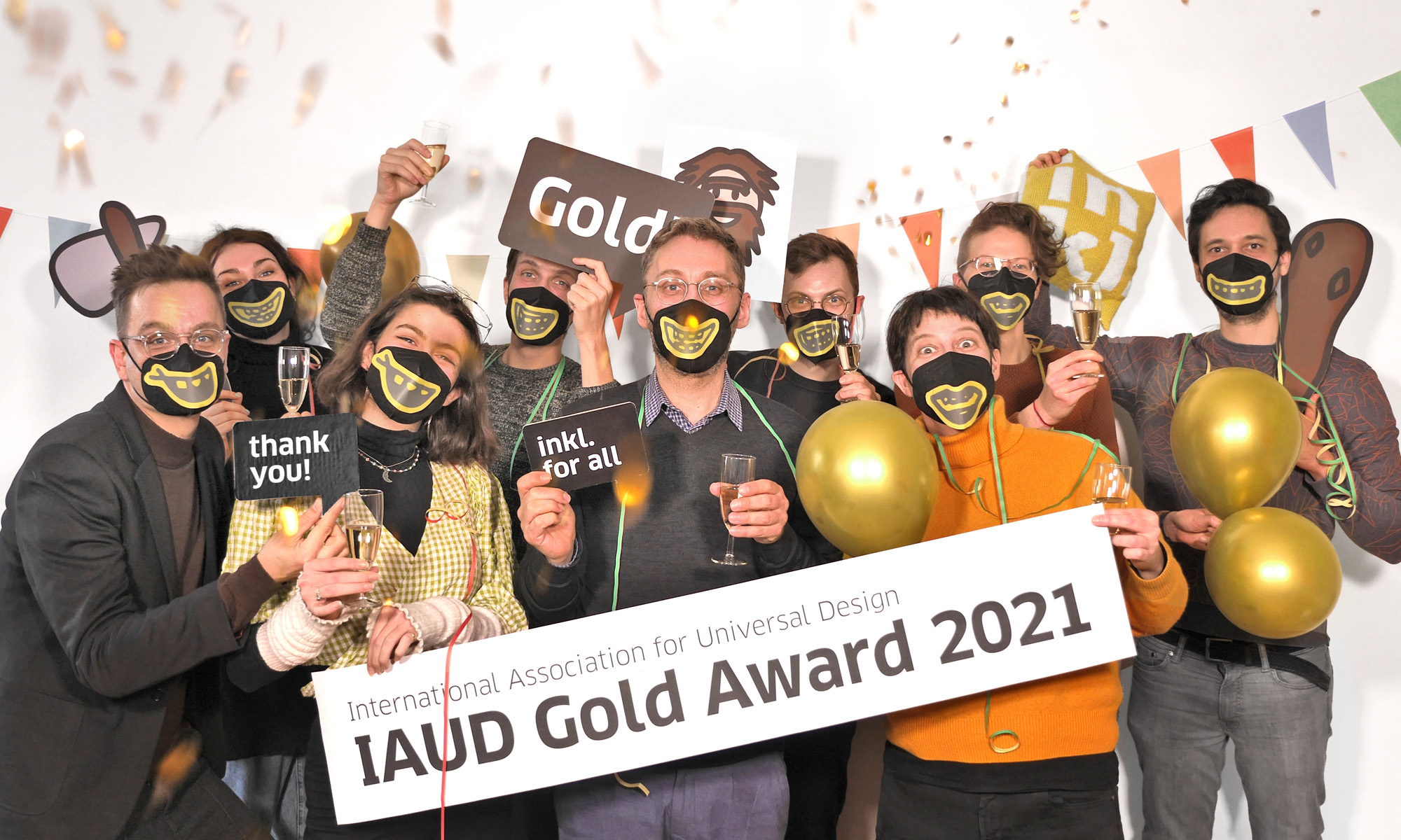 inkl-Mitarbeiterinnen und Mitarbeiter stehen Maske-tragend in Gruppe zusammen und freuen sich. Sie haben Sektgläser, Luftballons in der Hand, sind mit Girlanden behangen, von oben fällt Konfetti. Zwei Kolleginnen in der ersten Reihe halten ein großes Banner. Auf ihm steht: International Association for Universal Design, IAUD Gold Award 2021.