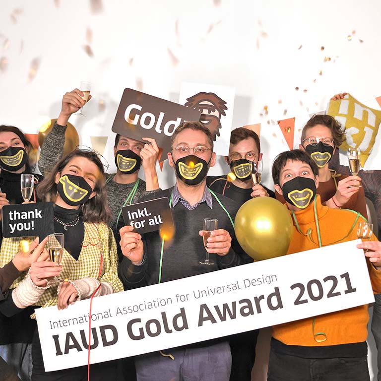 inkl-Mitarbeiterinnen und Mitarbeiter stehen Maske-tragend in Gruppe zusammen und freuen sich. Sie haben Sektgläser, Luftballons in der Hand, sind mit Girlanden behangen, von oben fällt Konfetti. Zwei Kolleginnen in der ersten Reihe halten ein großes Banner. Auf ihm steht: International Association for Universal Design, IAUD Gold Award 2021.