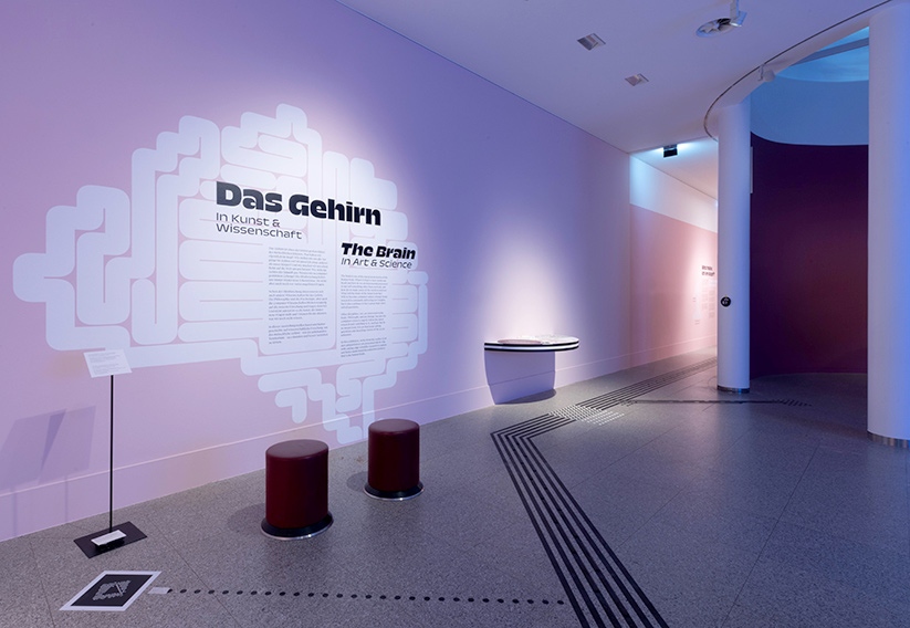 View into the entrance area of the exhibition „The Brain“ in the Bundeskunsthalle Bonn. On the left on the pink wall, a graphic representation of the brain with information texts. In front of it, two round stools. The floor guidance system leads past the first inclusive table into other areas of the exhibition.
