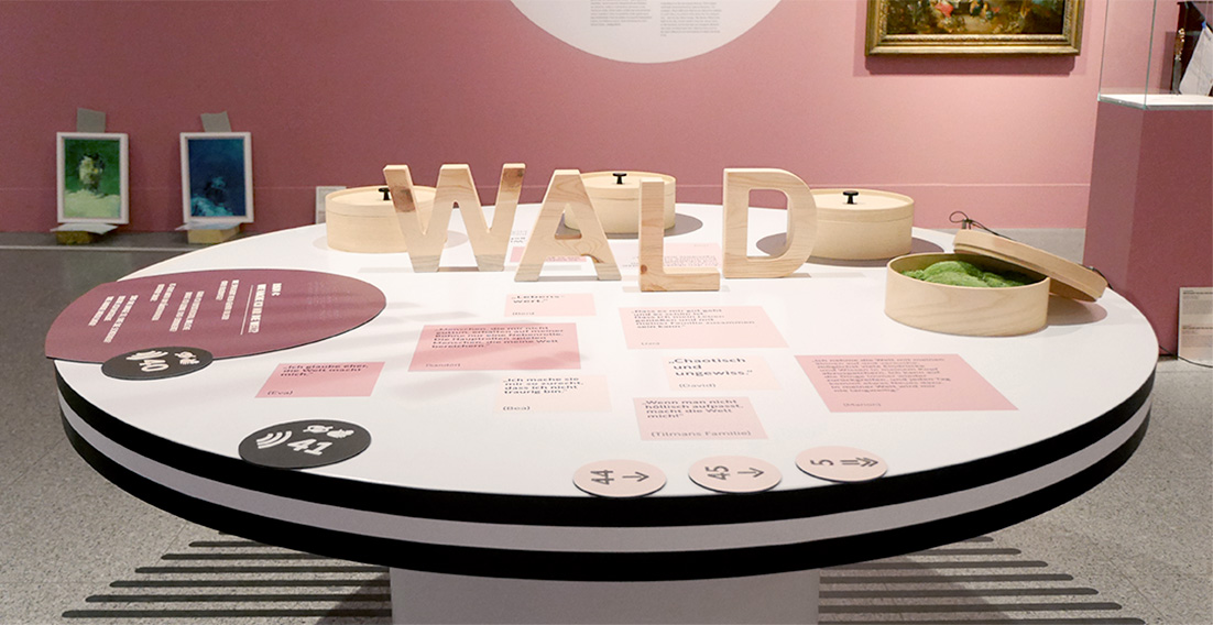 Photo of an inclusive table. In the center are large wooden letters that form the word „Wald“ Also, several round wooden boxes, some tactile signs and colored stickers. In one box the lid is slightly open, green moss is visible inside.