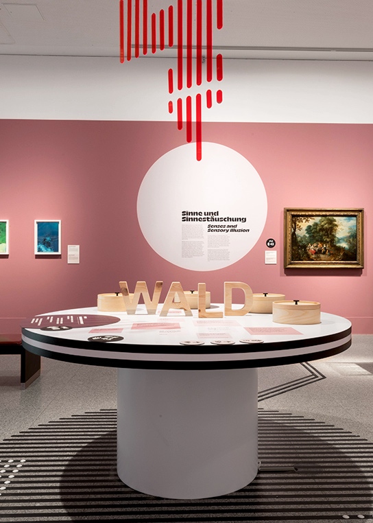 Another photo of an inclusive table. In the center of the table are placed letters made of wood, which make the word „Wald“. Also, round boxes made of wood, tactile signs and stickers. In the background a pink exhibition wall with text and two pictures.