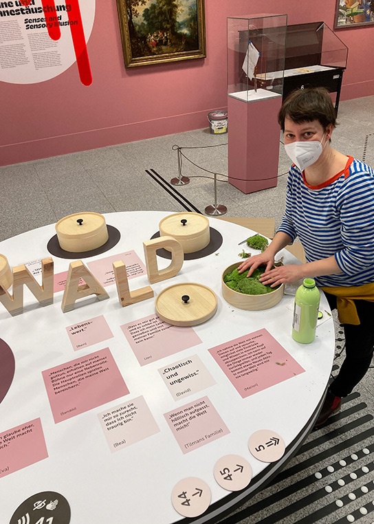 Foto der inkl.Design-Mitarbeiterin Franziska Müller, mit Maske, beim Aufbau. Mit den Händen berührt sie das Moos in einer Holzdose.