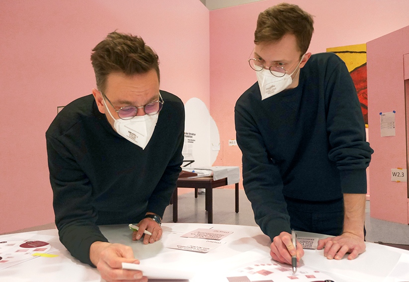 Zwei Mitarbeiter von inkl.Design bei der Montage von Ausstellungstischen in der Bundeskunsthalle Bonn. Sie stehen nebeneinander an einem der Tische und schauen konzentriert. Beide tragen eine Brille, Maske und dunkle Pullover.