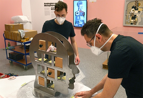 Das Foto zeigt Ludwig und Gregor von inkl.Design beim Aufbau der inklusiven Tische in Bonn. Zwischen ihnen ein großer Setzkasten aus Metall in Form eines Kopfes. Im Hintergrund ein Wagen mit Werkzeug.