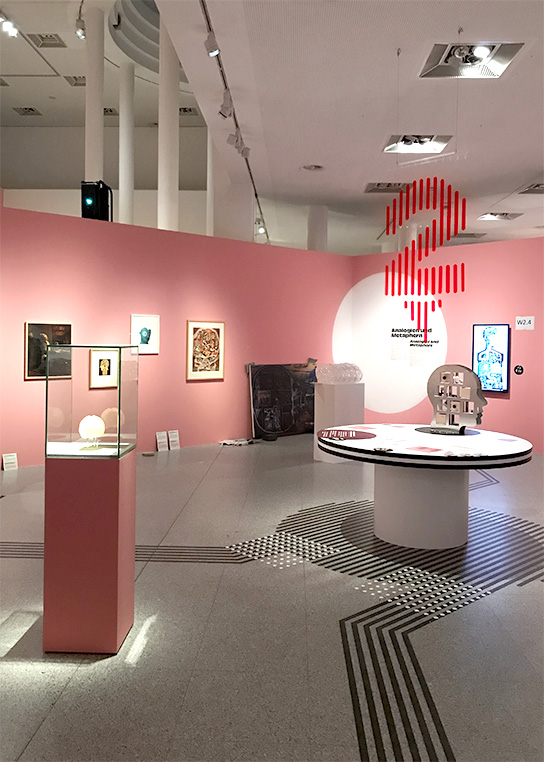 The picture shows the inclusive table number 2 and the surrounding exhibition. On the table the large set box in the shape of a head. The distinctive floor guidance system leads to the table and other areas of the exhibition.