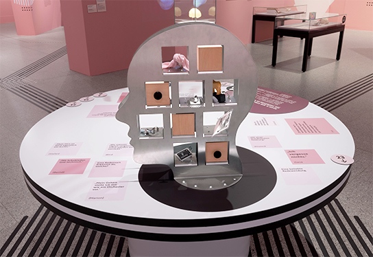 The photo shows the entire inclusive table 2 with the set box in the shape of a large head. On the table surface you can see stickers in different shades of pink with short quotes from the focus group and tactile marks for orientation.
