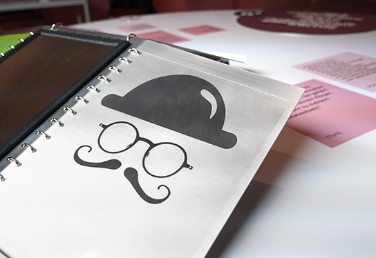 Close-up of an exhibition object on one of the inclusive tables: a transparent film on which a round hat, glasses and a curved mustache are printed in black lie above a mirror.