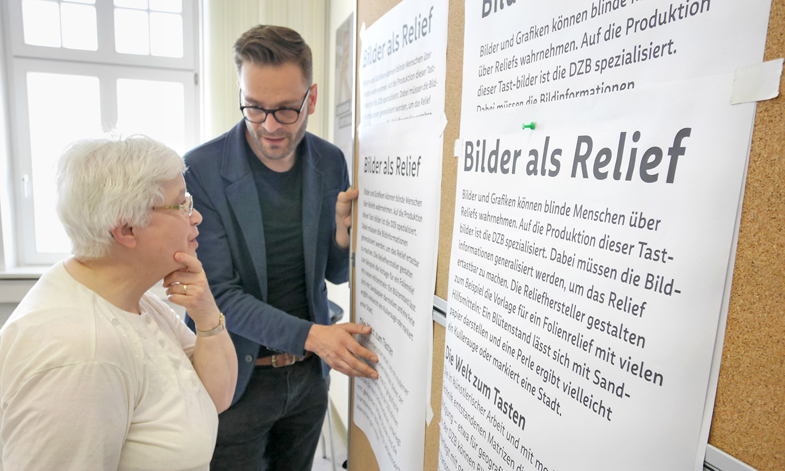 Photo from a focus group meeting on font legibility. Gregor Strutz in conversation with an elderly woman from the focus group. They are standing together in front of a large board with text pinned to it in different font sizes.
