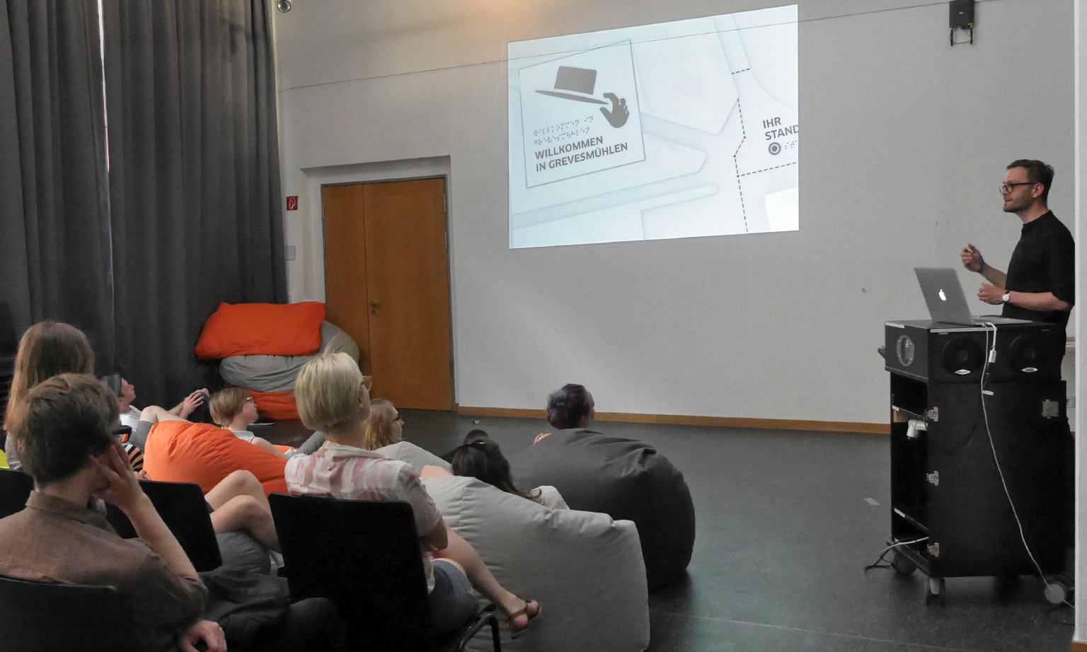 Photo of a lecture at the University of Applied Sciences in Berlin. On the right of the picture, Gregor Strutz stands at a lectern and speaks to the audience. Students sitting on chairs and beanbags are looking with interest at a beamer projection on the wall. The room is slightly darkened and on the wall you can see a section of a tactile plan with focus on an icon and Braille.